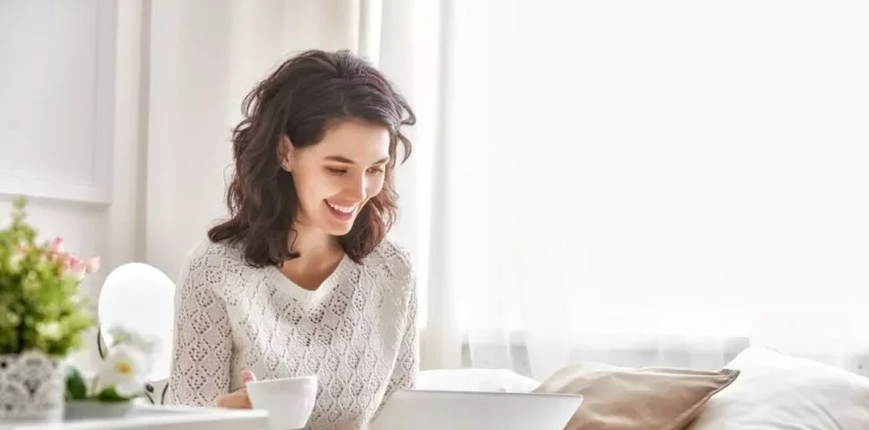 Person smiling while using a laptop in a bright room.