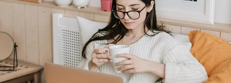 Person on a couch sipping coffee while using a laptop.