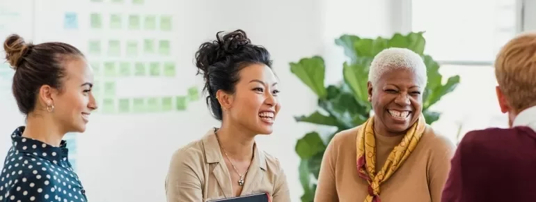 Four women are smiling and engaged in conversation in a bright office setting.