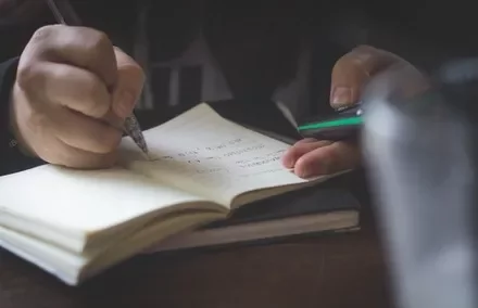 A pair of hands writing in a notebook while holding a phone.