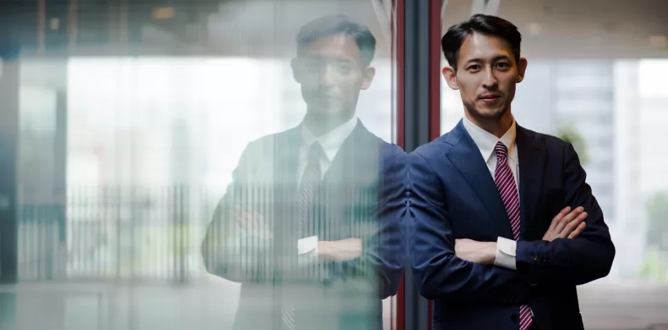 A person in a suit leaning on a glass wall with folded arms.